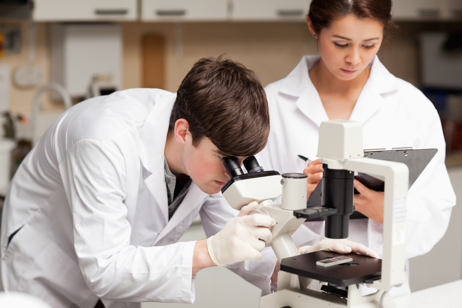 Male scientist looking in a microscope while his female colleague is taking notes.