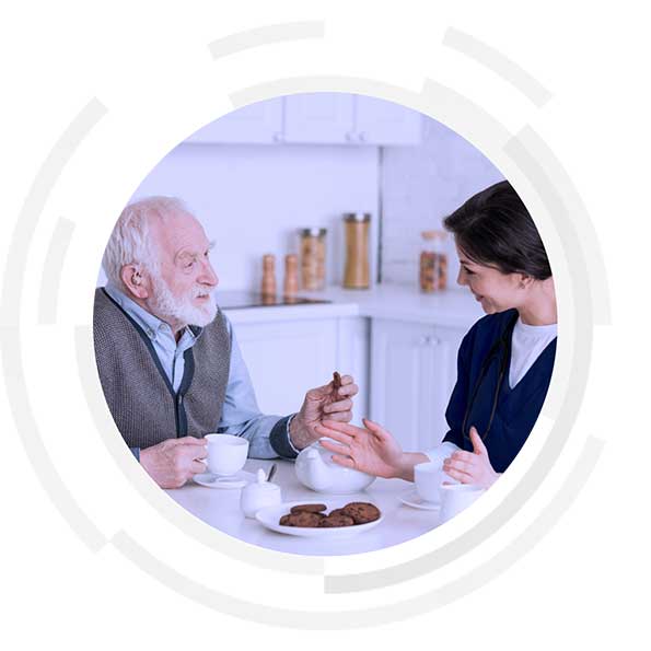 Elderly patient and nurse sitting at dining table sharing tea and biscuits.
